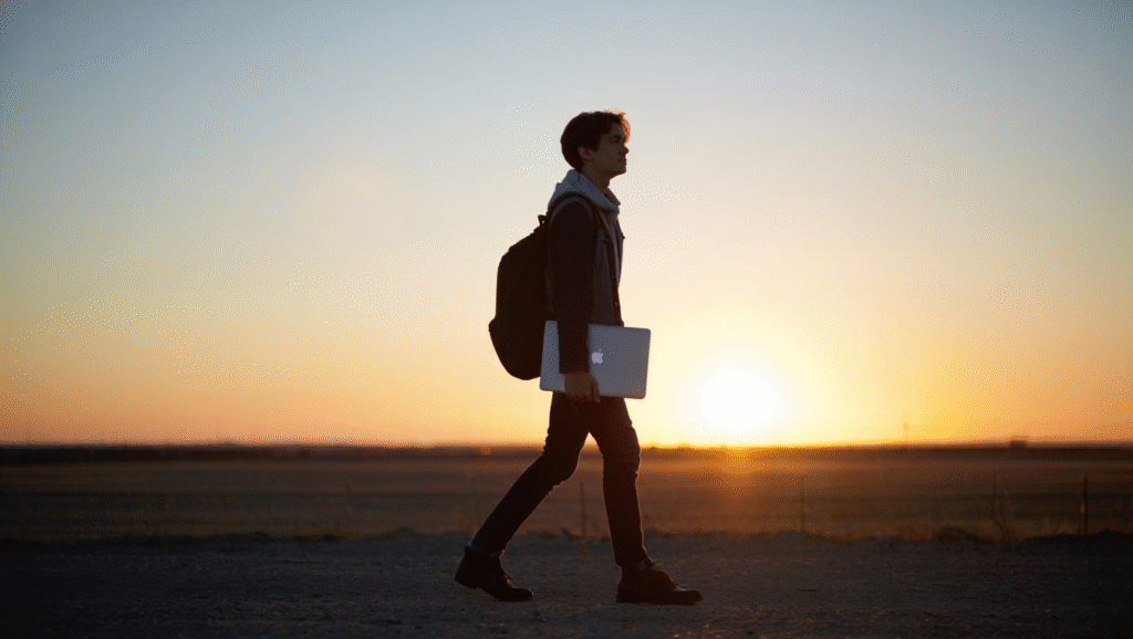 La Mentalità del Costruttore Giovane cammina al tramonto con MacBook e zaino verso libertà professionale - simbolo del percorso da zero a uno nell'economia dei creator